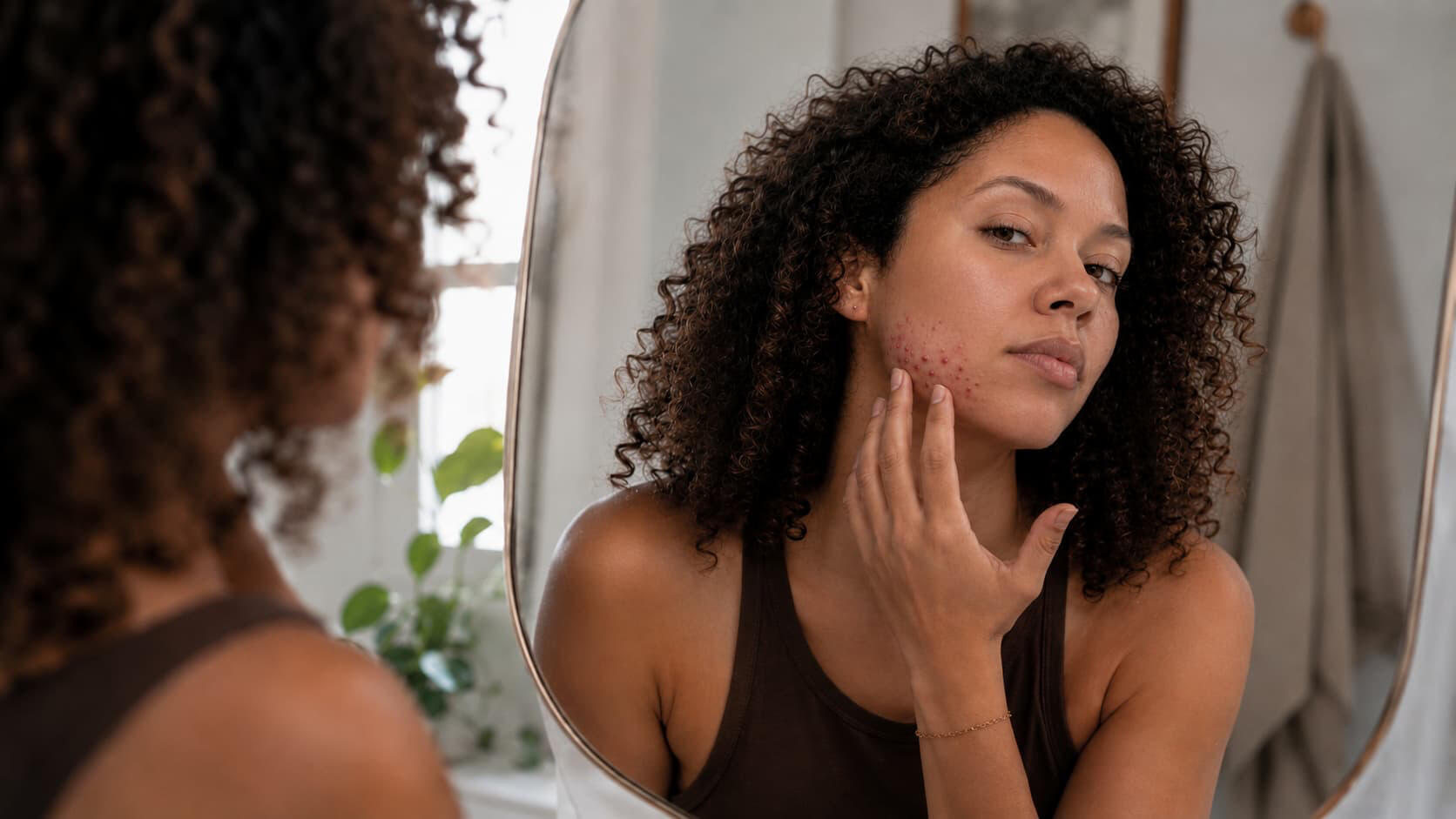 Adult woman in her 30s looking at her reflection in a bathroom mirror, examining recurring breakouts on her jawline that return in the same spot every month